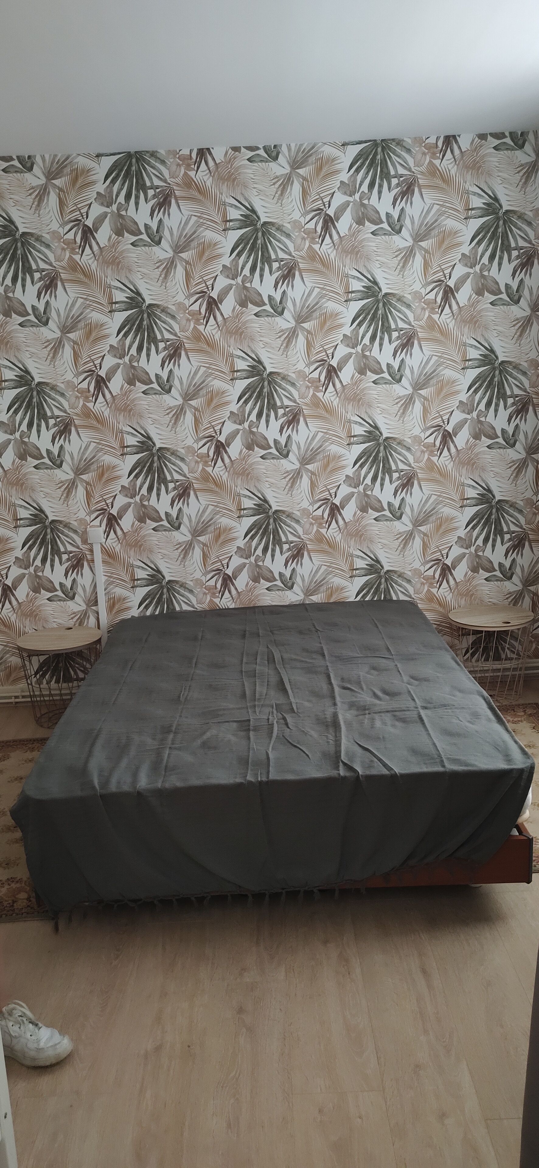 Habitació amb un llit cobert per un llençol gris, un paper pintat i una taula petita rodona amb una cadira.