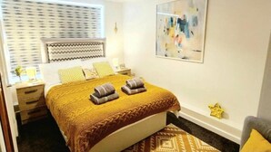 Ein ordentlich eingerichtetes Schlafzimmer mit Bett, Nachttischen, einer Nachttischlampe, einem Fenster mit Jalousien und einem an der Wand hängenden abstrakten Gemälde.
