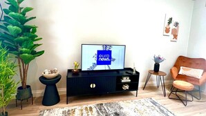Un salon avec un téléviseur diffusant les informations européennes, un meuble de couleur sombre, une table basse ronde en bois, une petite table d’appoint, une chaise et plusieurs plantes en pot.