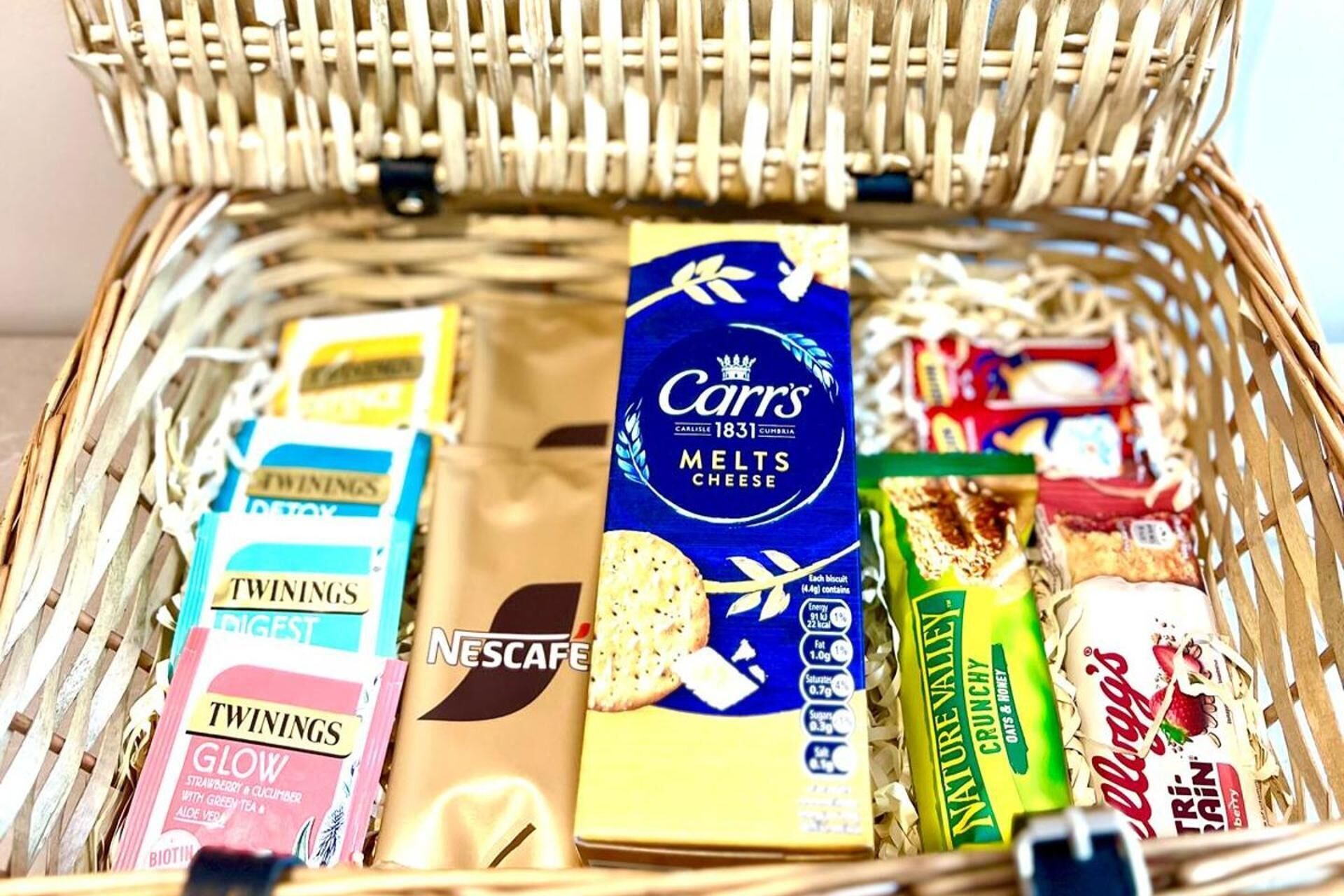 A wicker basket filled with various food items including a box of Carr's Melted Cheese, Twinings tea, Nescafe, and other snacks.