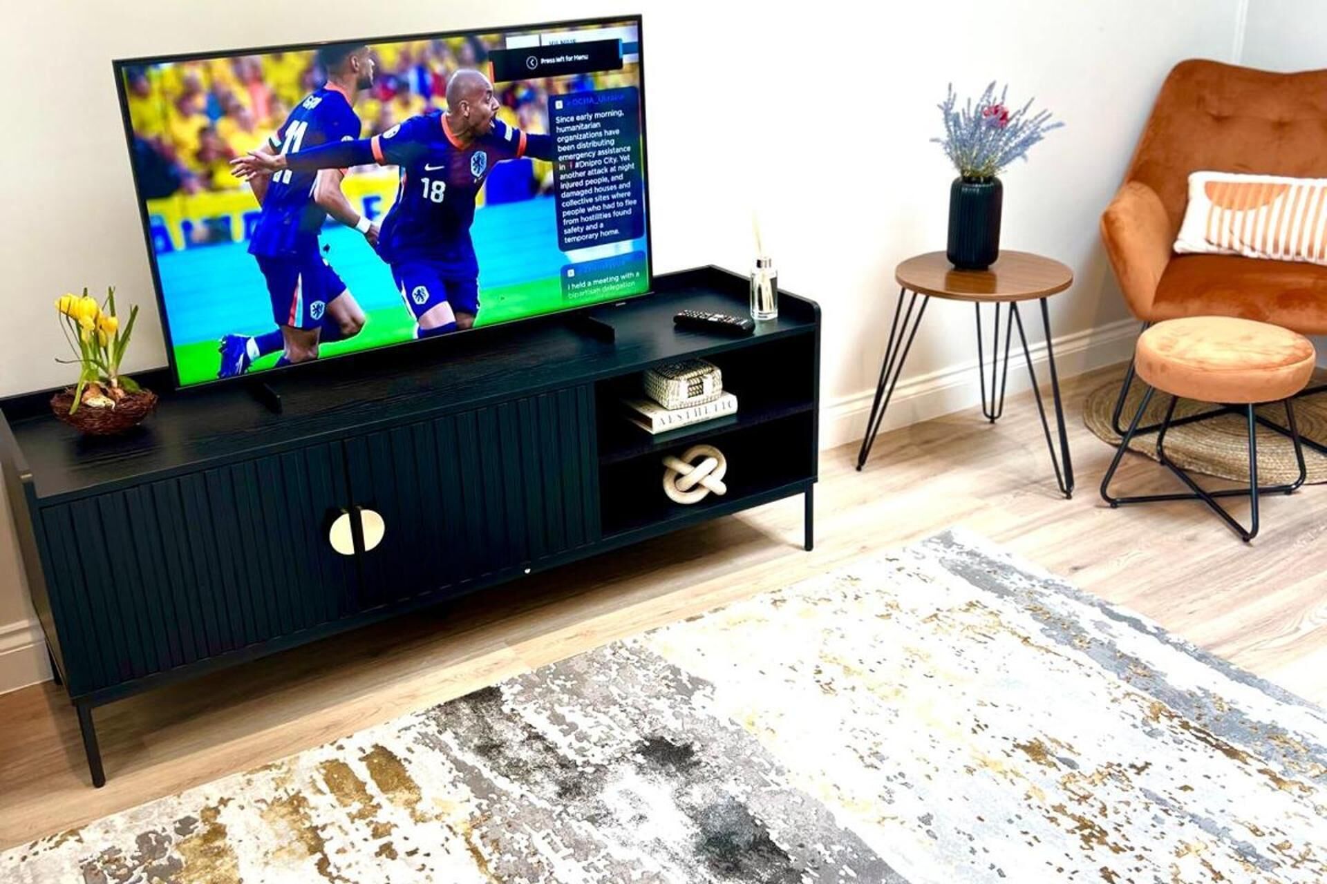 A modern living room with a dark wooden entertainment center, a flat-screen TV displaying a sports event, a small side table with a vase, and a cozy orange chair.