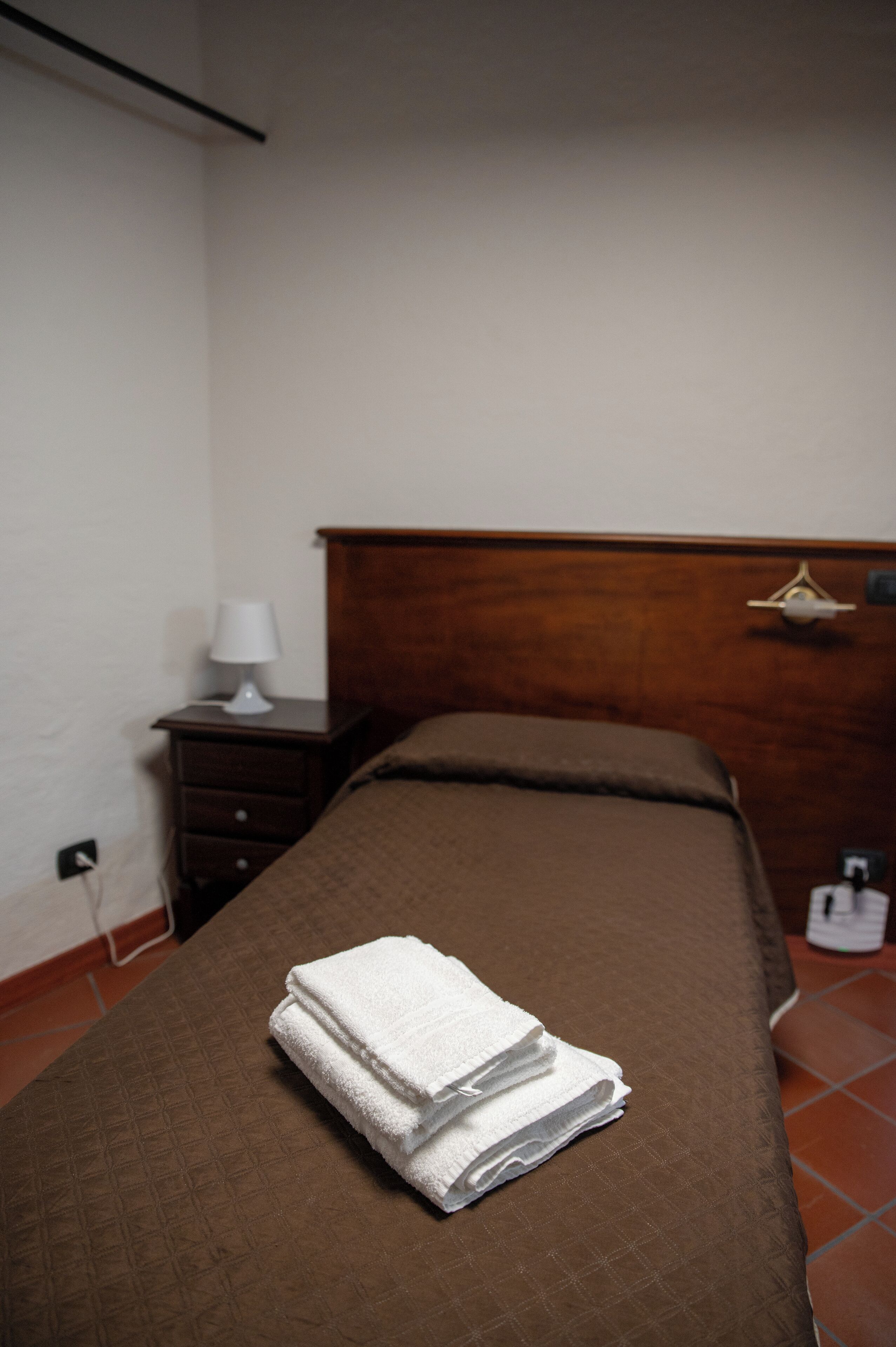 Un llit ben arrenjat amb un estovat marró i tovalloles blanques doblegades a sobre. Hi ha una nitòria de fusta amb una làmpada blanca al costat del llit.