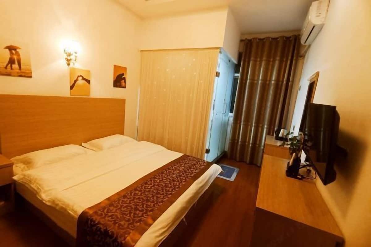 A hotel room with a bed, a nightstand, a desk with a chair, a wall-mounted TV, and a window with curtains.