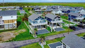 A coastal residential area with elevated houses featuring balconies and decks, surrounded by green fields and a body of water.