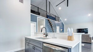 A modern kitchen with a central island, stainless steel dishwasher, and a black staircase leading to an upper level.