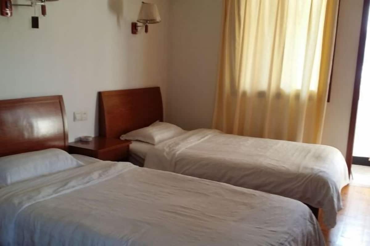 A hotel room with two single beds, a wooden headboard, a nightstand, a lamp, and a window with curtains.