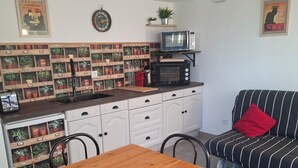 A compact living space with a kitchenette, a dining table with four chairs, a striped sofa with a red pillow, and a wall-mounted plant shelf. - Gite Cosy Dans un Cadre Verdoyant Entre Méditerranée et Gorges du Verdon (Flassans-sur-Issole)