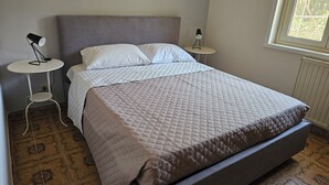 A neatly made bed with a quilted bedspread and pillows, a small round table with a lamp, and a window with a view of greenery.