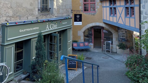 Una calle estrecha con edificios de arquitectura tradicional, incluyendo un café con terraza y un cartel que dice 'Le Café Canine'.