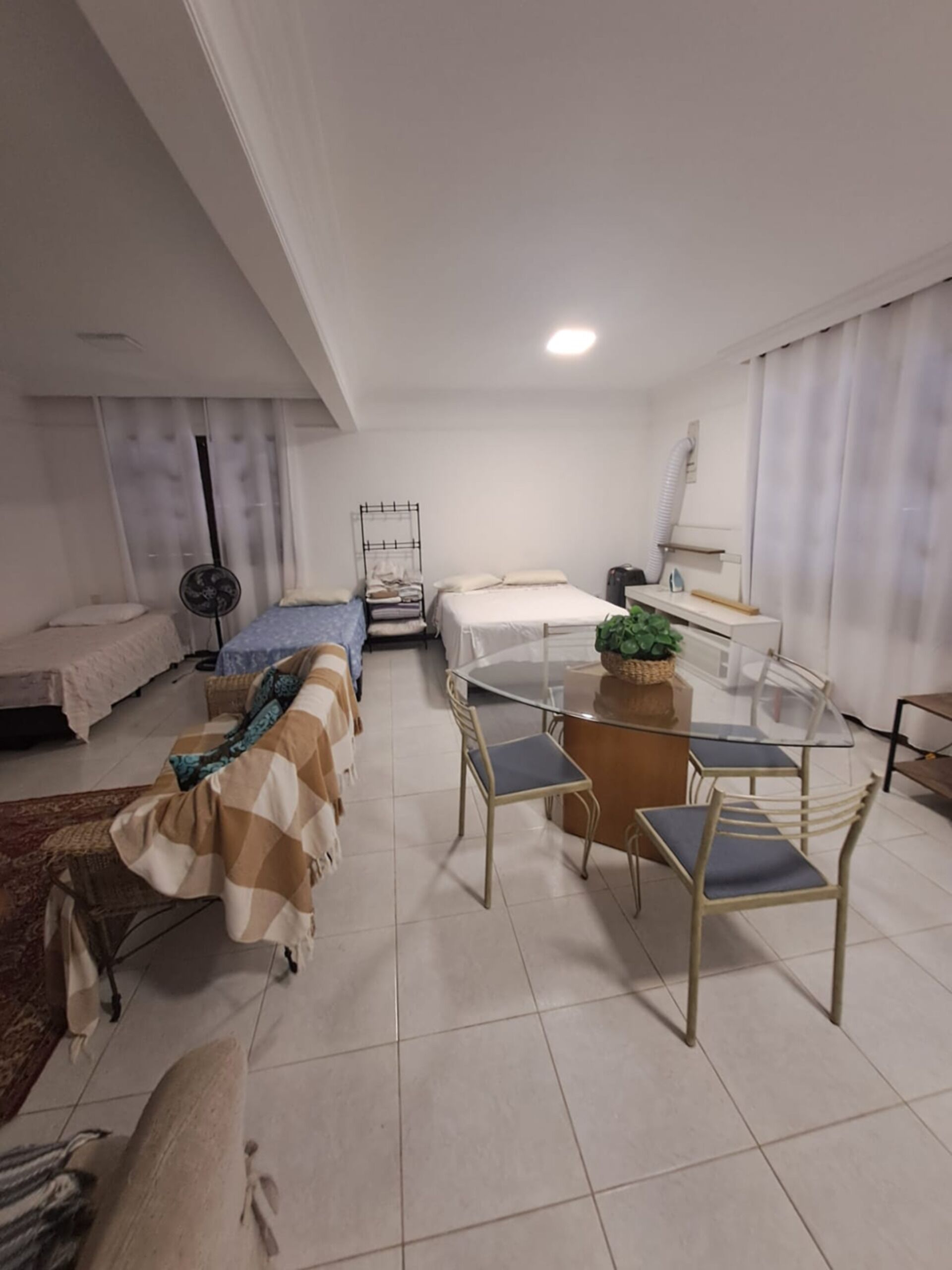 Una habitación de hotel moderna con cama, sofá, mesa de comedor con sillas, un ventilador de pie y una planta pequeña.