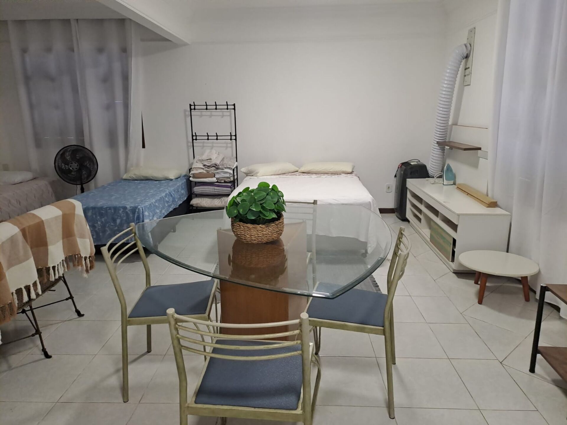 A compact hotel room with a glass dining table, two chairs, a bed with a blue bedspread, a small sofa, a standing heater, a fan, and a shelf with folded clothes.