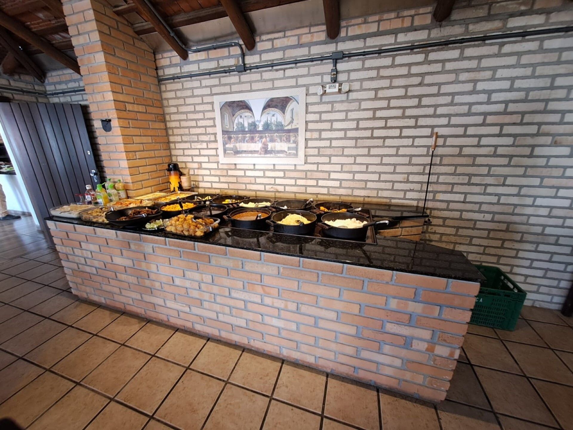 A buffet setup with various dishes on a brick counter, a tiled floor, and a brick wall with a framed picture.