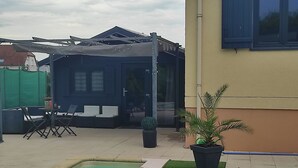 Un área de piscina con una zona de estar cubierta, una pequeña mesa con sillas y una planta en maceta.
