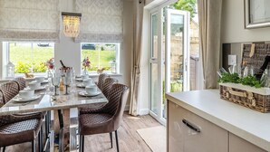 Un coin repas avec une table dressée pour quatre personnes, deux chaises rembourrées, un luminaire suspendu et une vue sur un jardin grâce à de grandes fenêtres.