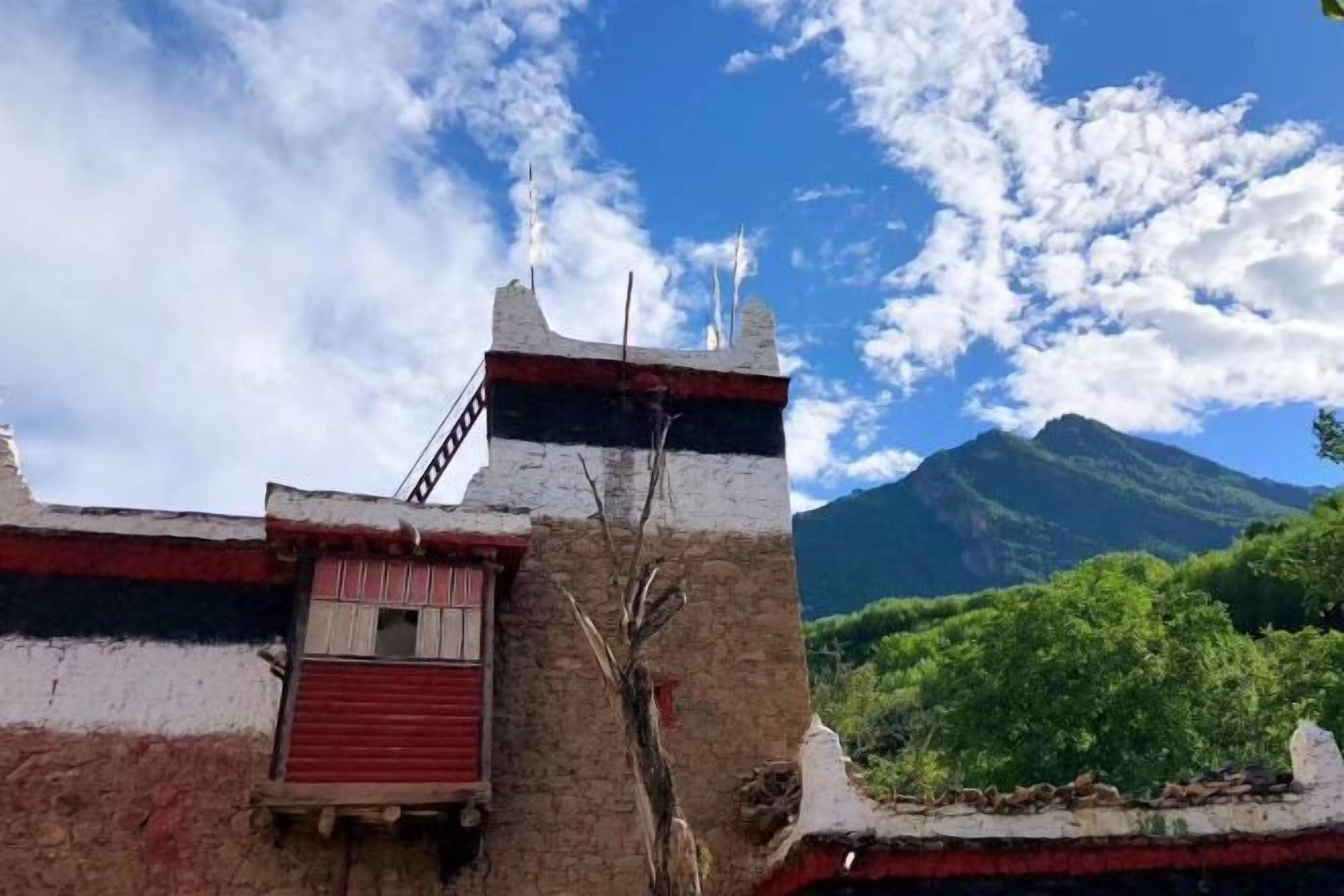 Un bâtiment avec une fenêtre à volets rouges et une cheminée, se dressant sur fond de montagne et d’un ciel partiellement nuageux.
