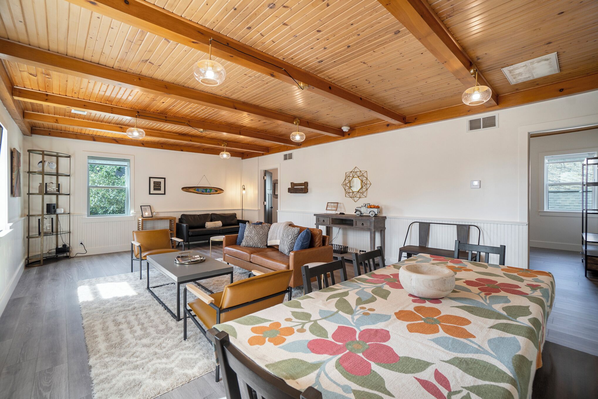 A spacious living room with a high ceiling, wooden beams, a large dining table with a floral tablecloth, and a cozy seating area with a sofa and chairs.