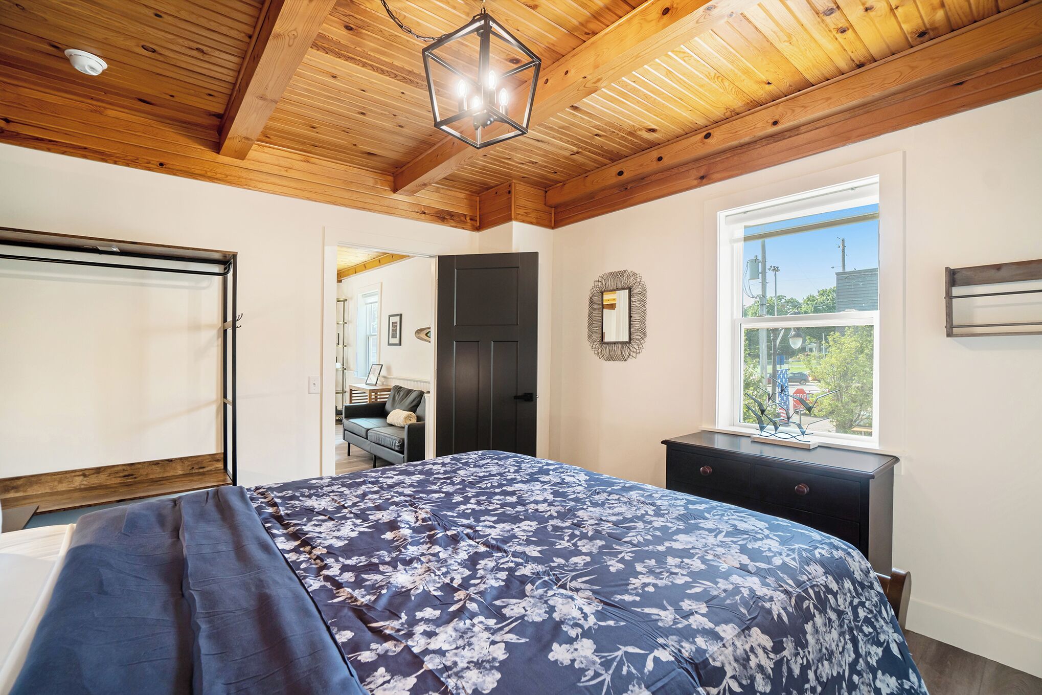 Une chambre avec un plafond en bois, une grande fenêtre, un lit recouvert d’une couvre-lit à motifs floraux et une commode.