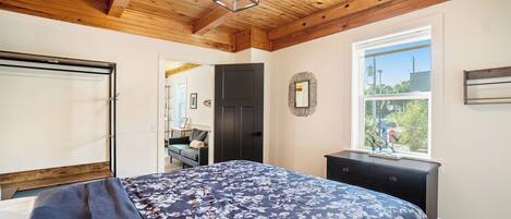 Une chambre avec un plafond en bois, une grande fenêtre, un lit recouvert d’une couvre-lit à motifs floraux et une commode.