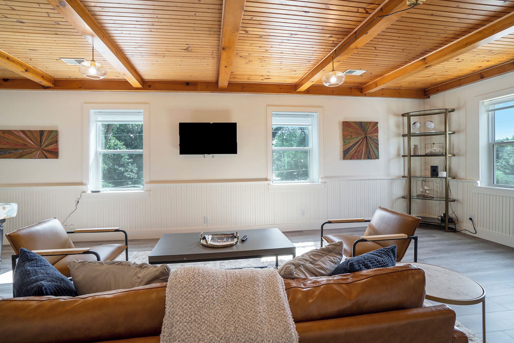 A spacious living room with a wooden ceiling, a flat-screen TV mounted on the wall, two windows, a sofa, and a coffee table.