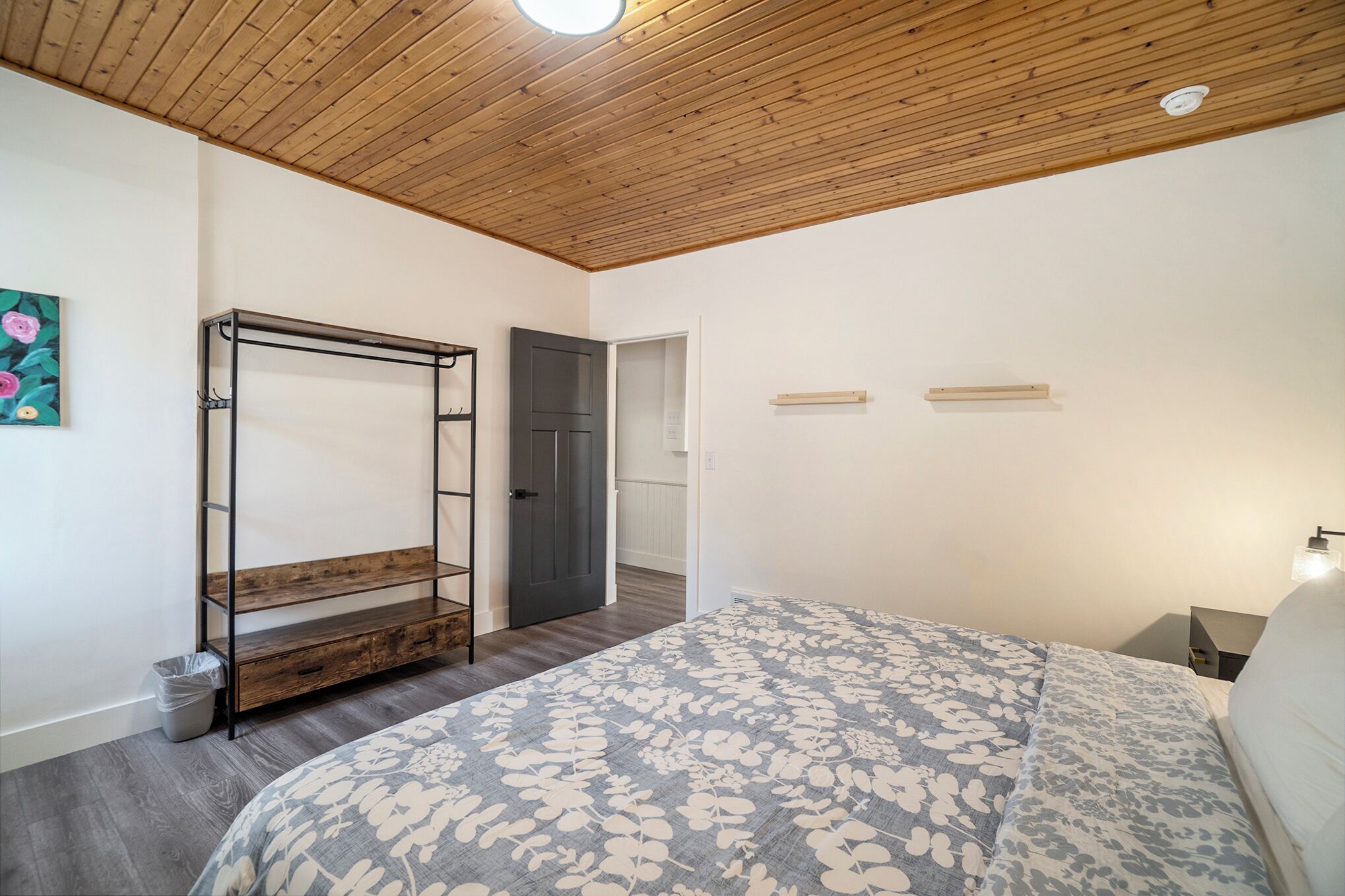 A bedroom with a wooden ceiling, a bed with a floral bedspread, a metal wardrobe, and a door leading to another room.