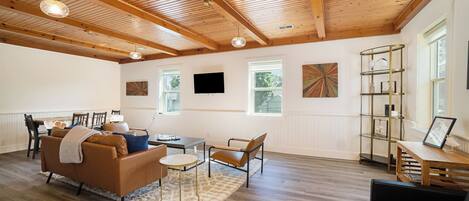 A spacious living area with wooden beams, a fireplace, a dining table with chairs, a sofa, a coffee table, and a bookshelf.
