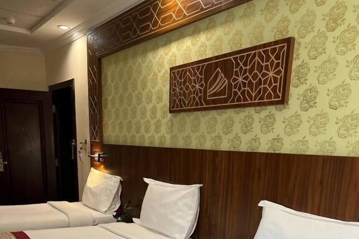A hotel room with two beds, each with white bedding and a red accent. There is a wooden headboard with a geometric design, and a wall-mounted decorative panel above the beds.