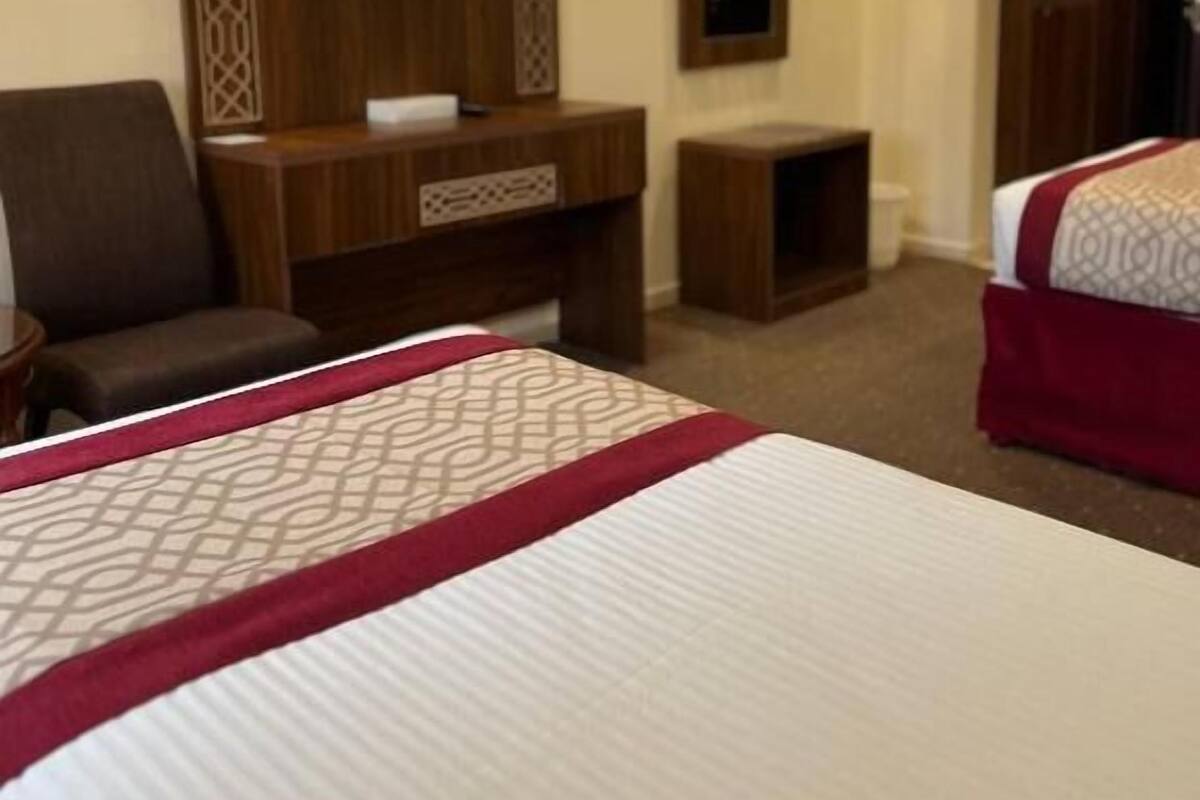 A hotel room with a large bed, a TV mounted on a wooden panel, a desk, a chair, and a wardrobe.