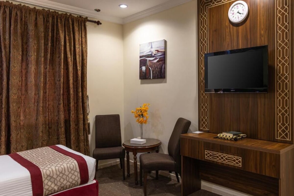 A hotel room with a bed, two chairs, a desk, a TV, and a clock on the wall.