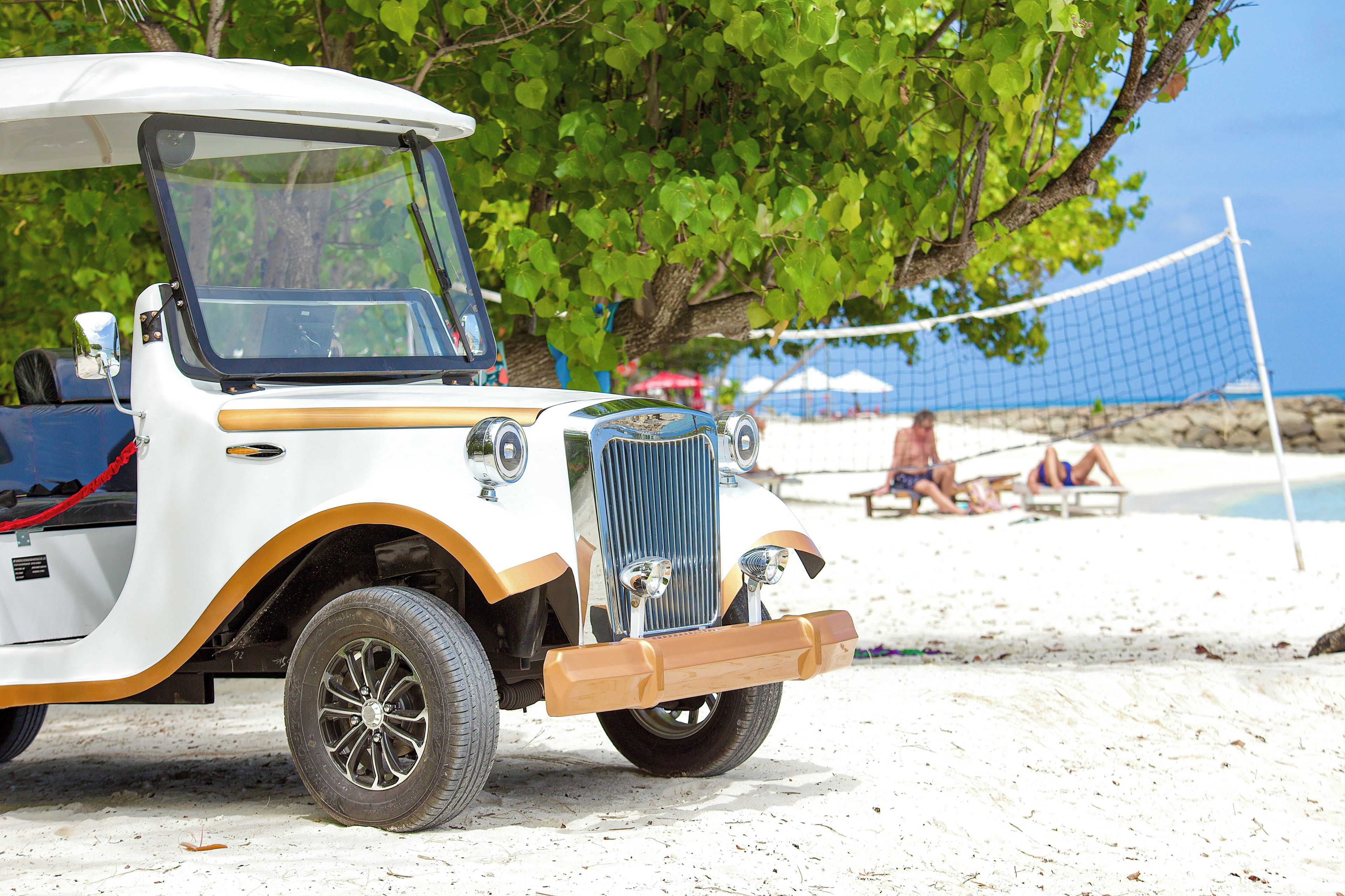 A vintage car parked on a sandy beach with a clear blue sky and a volleyball net in the background.