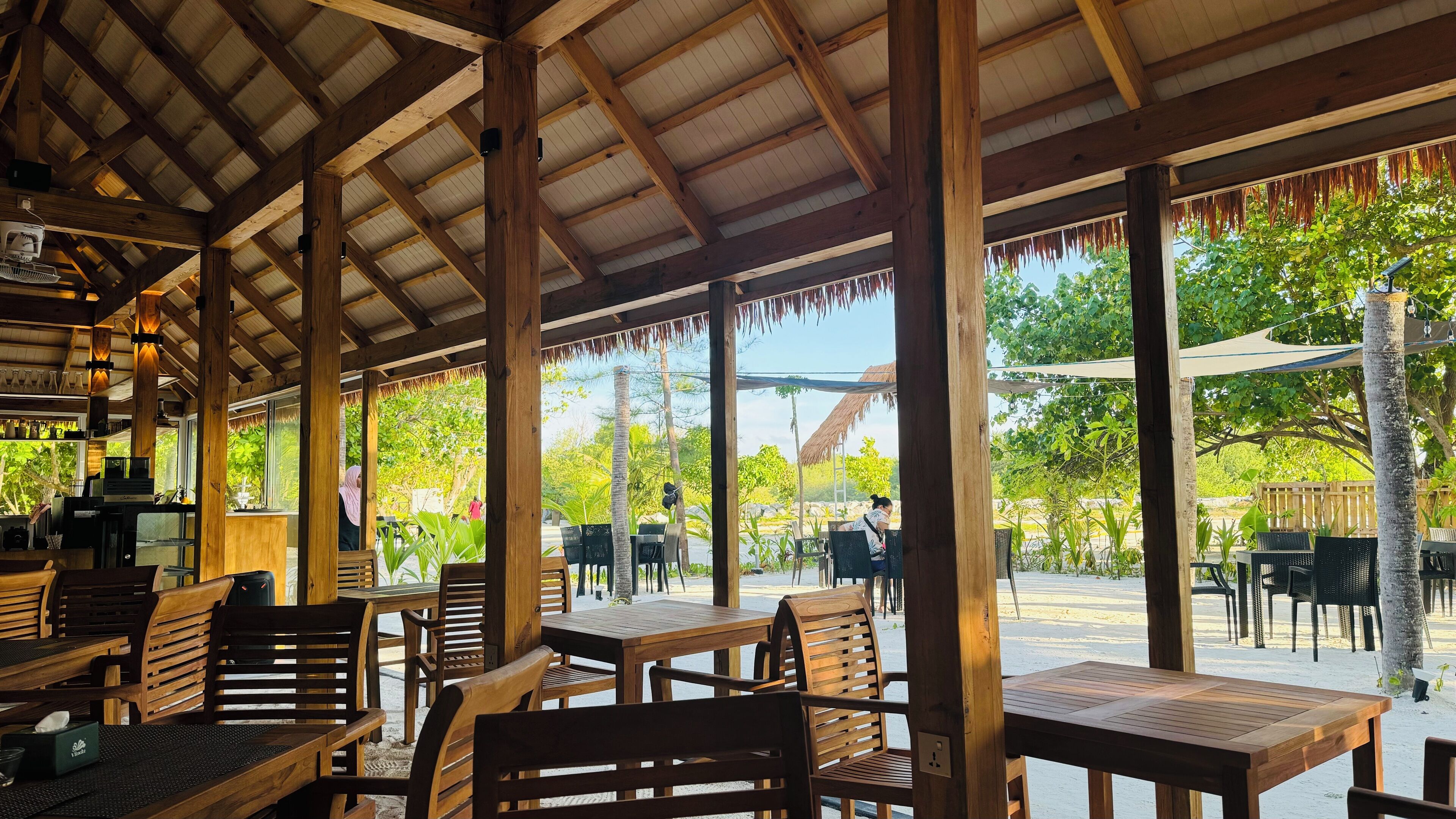 An outdoor restaurant with wooden tables and chairs, a covered roof, and a view of the surrounding greenery.