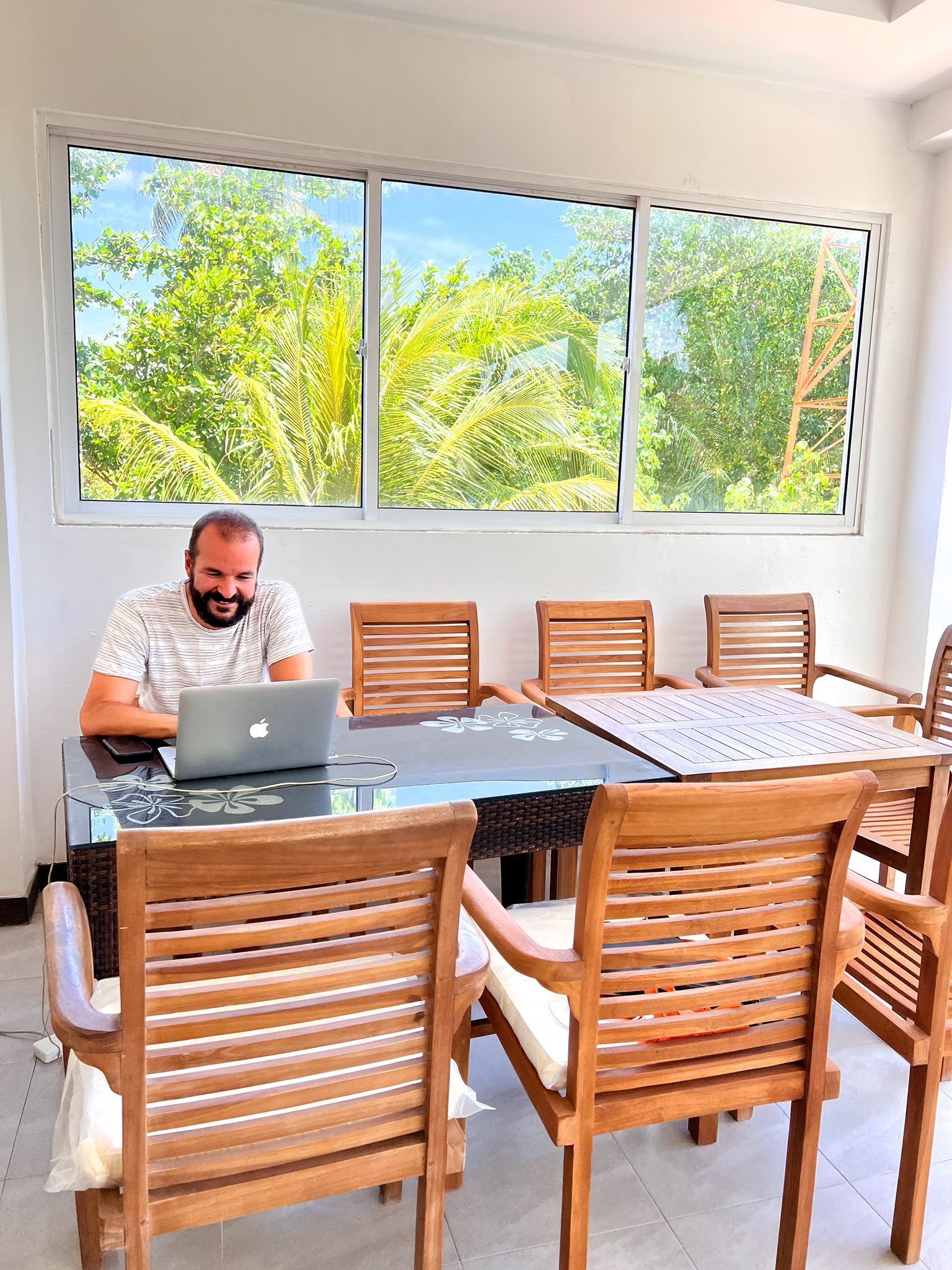 A person seated at a table with a laptop, surrounded by wooden chairs, in a room with large windows overlooking greenery.