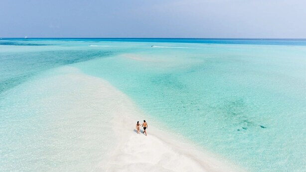Un couple se promène sur une plage de sable fin, bordée d’une eau turquoise et cristalline.
