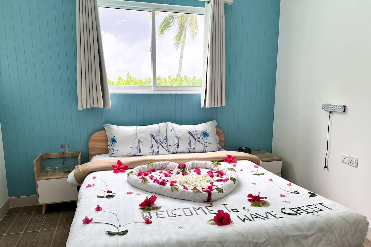 A bedroom with a bed, a bedside table, a nightstand, a window with curtains, and a wall-mounted power outlet.