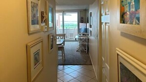 A hallway with framed pictures on the walls, a dining area with a table and chairs, and a view through a glass door.