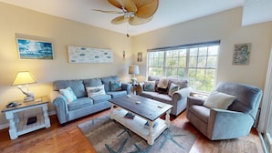A spacious living room with a blue sofa, a wooden coffee table, and a grey armchair. There are two side tables with lamps, a ceiling fan, and artwork on the walls. - Spacious, Marsh View Getaway w/ Multiple Outdoor Decks (Tybee Island)
