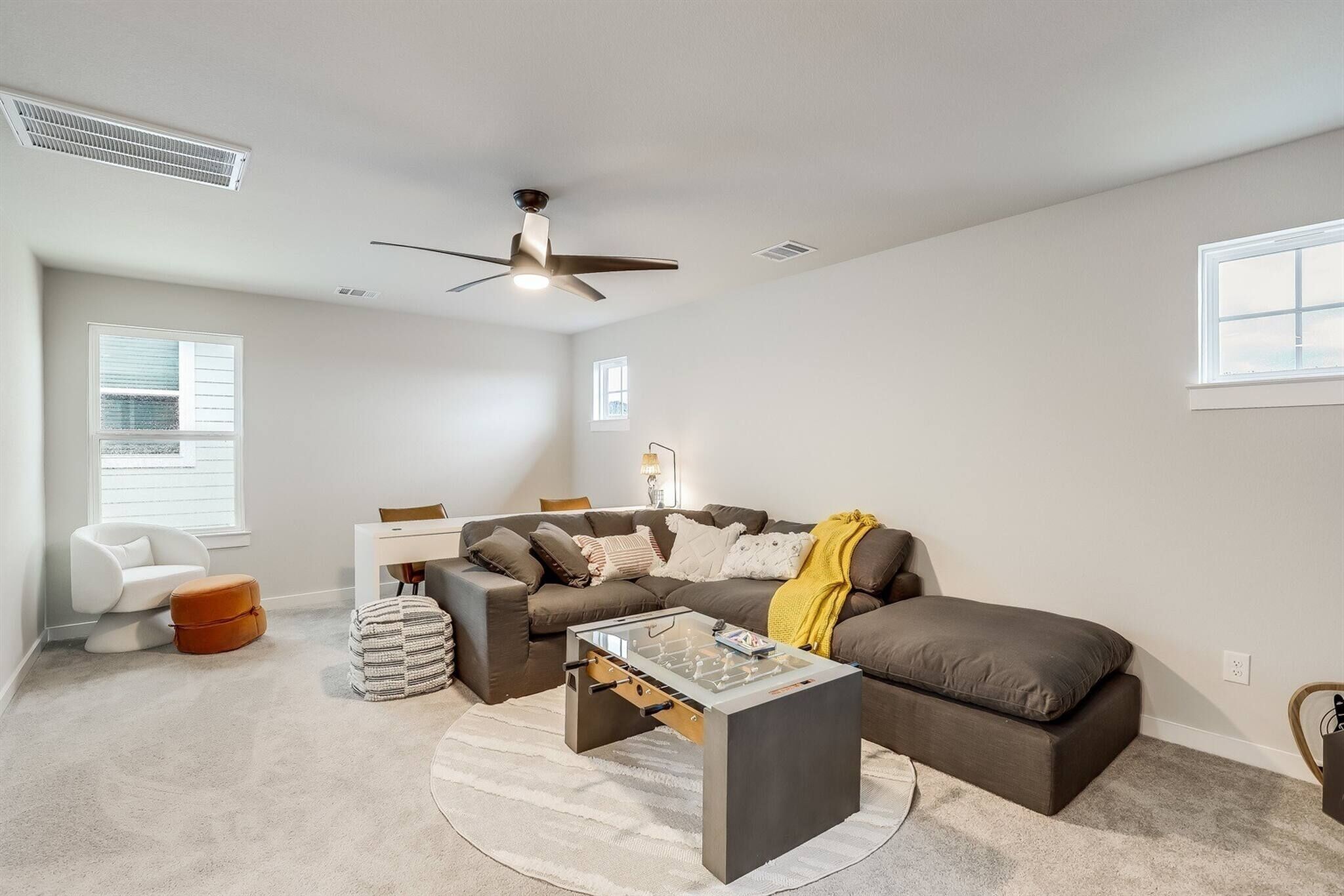 Un salon moderne avec un canapé d’angle, une table basse en verre, un ventilateur de plafond et une fenêtre équipée de stores.