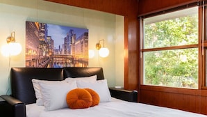 Une chambre d’hôtel avec un grand lit, deux lampes fixées au mur, un tableau représentant un paysage urbain et une fenêtre donnant sur un espace verdoyant.