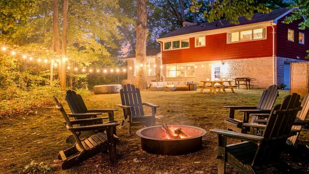 Un espace extérieur accueillant, aménagé avec des fauteuils Adirondack et un foyer, entouré d’une terrasse en bois et illuminé de guirlandes lumineuses.