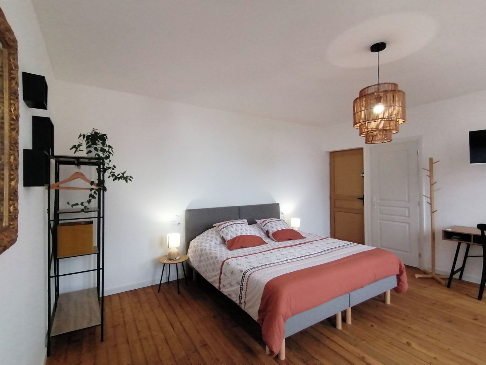 Une chambre à coucher avec un lit, des tables de chevet, une plante suspendue, un plancher en bois, un luminaire au plafond et une porte.