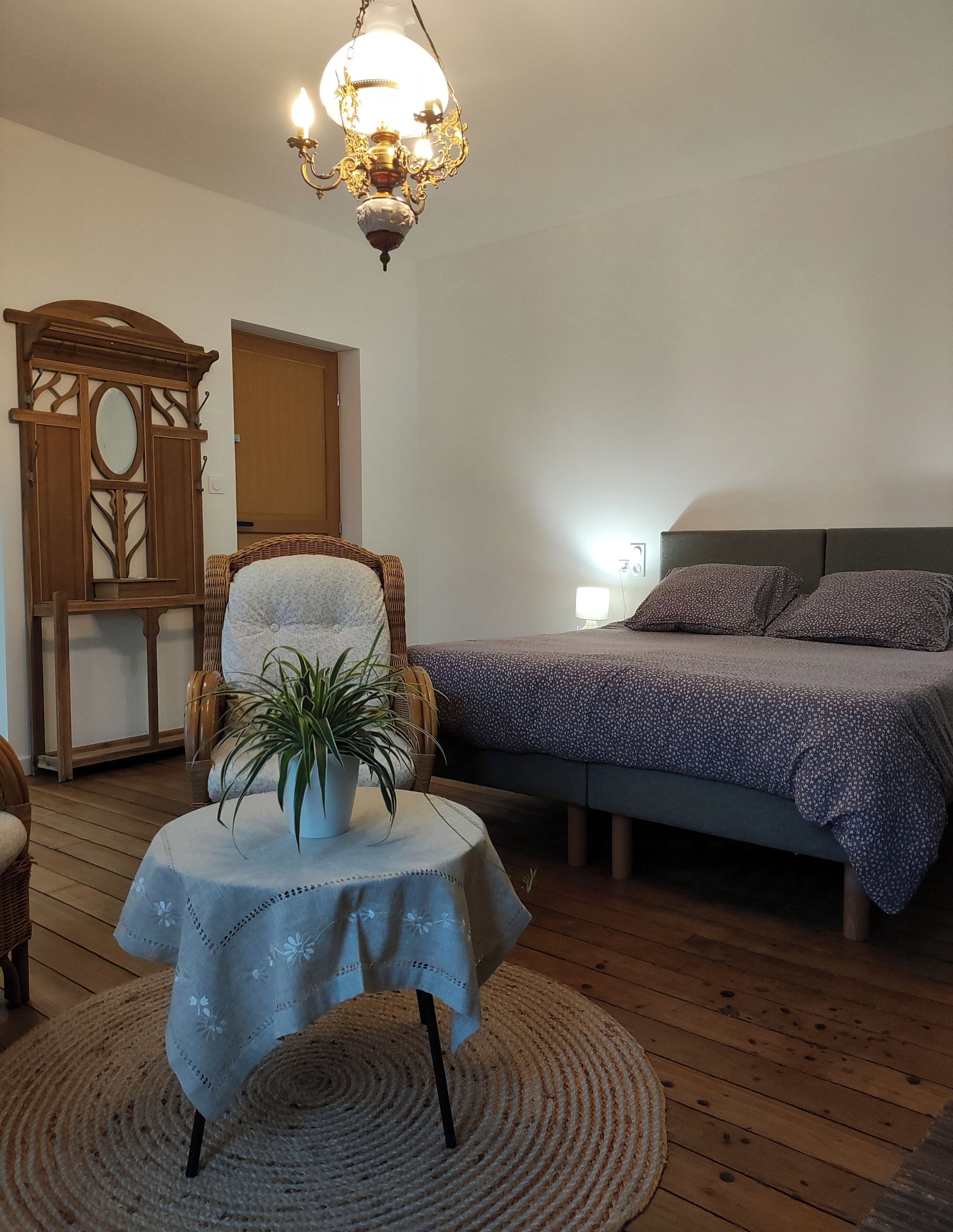 A cozy bedroom with a bed, a chair, a table with a plant, and a chandelier.