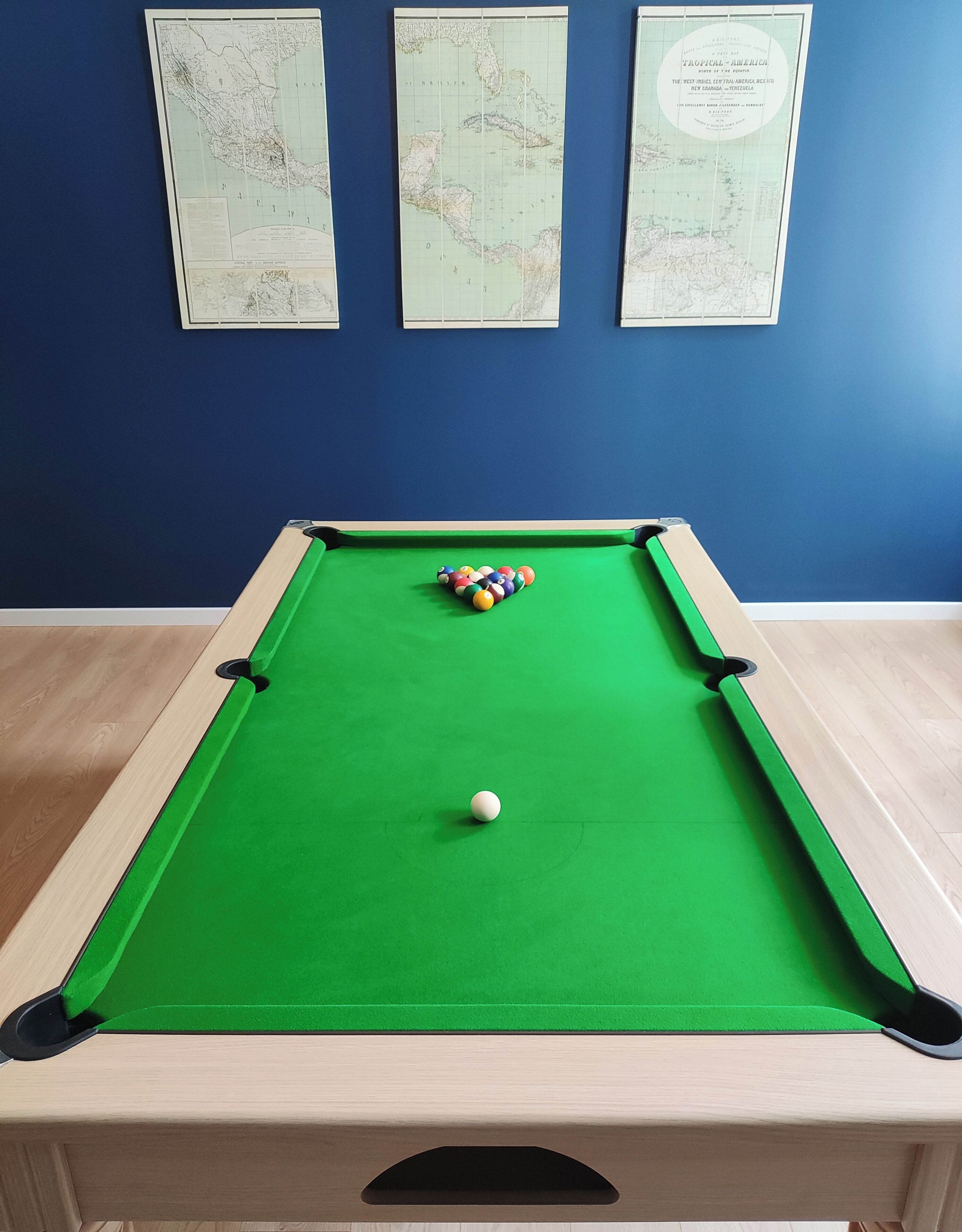 Uma mesa de bilhar com bola branca e bolas coloridas, ao fundo uma parede azul com três mapas.