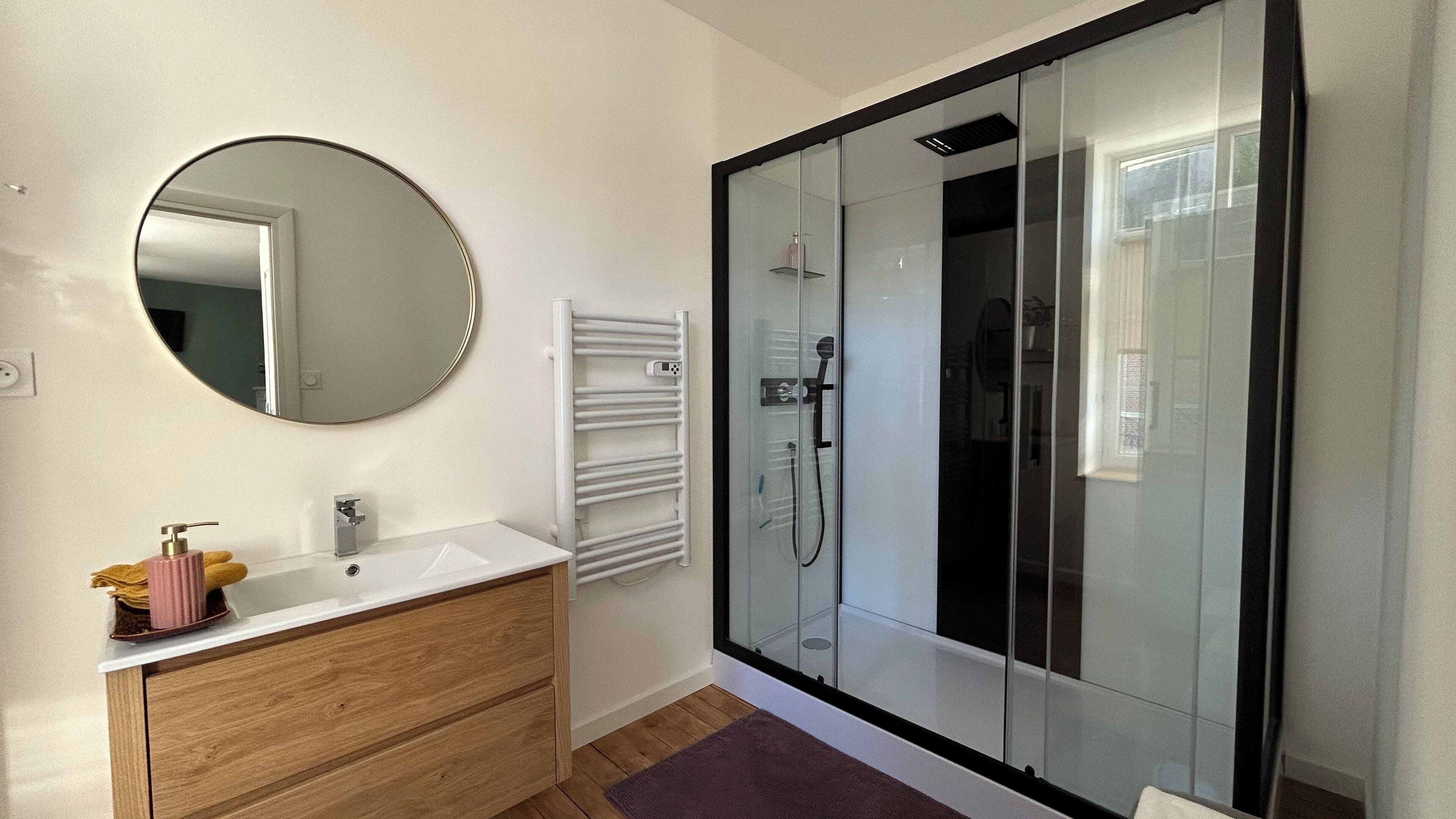 Un baño moderno con un espejo redondo, un lavabo blanco, un tocador de madera, una cabina de ducha de vidrio y un perchero para toallas.
