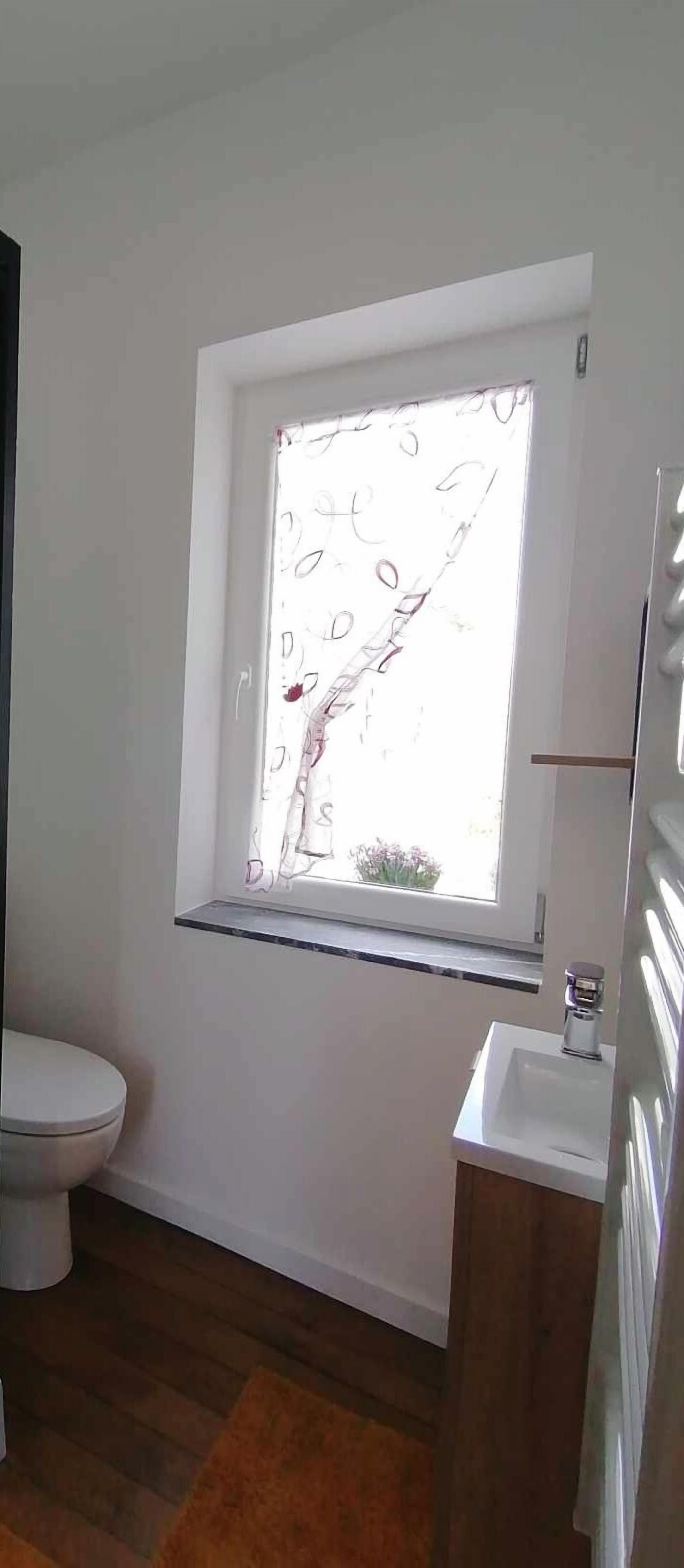 A bathroom with a toilet, a sink, a window with a view of the outside, and a wooden floor.