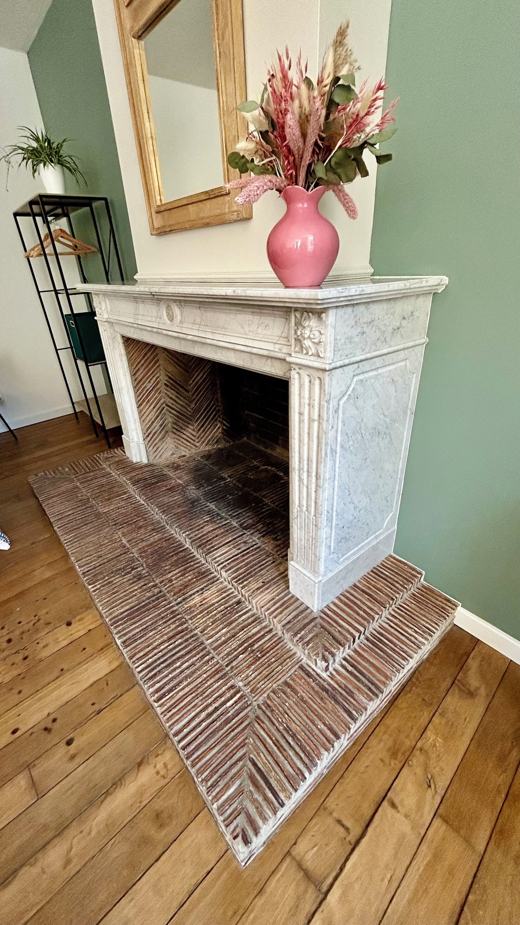 A fireplace with a pink vase of dried flowers on top, a patterned rug in front, and a wooden floor.
