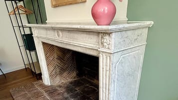 A fireplace with a pink vase of dried flowers on top, a patterned rug in front, and a wooden floor.