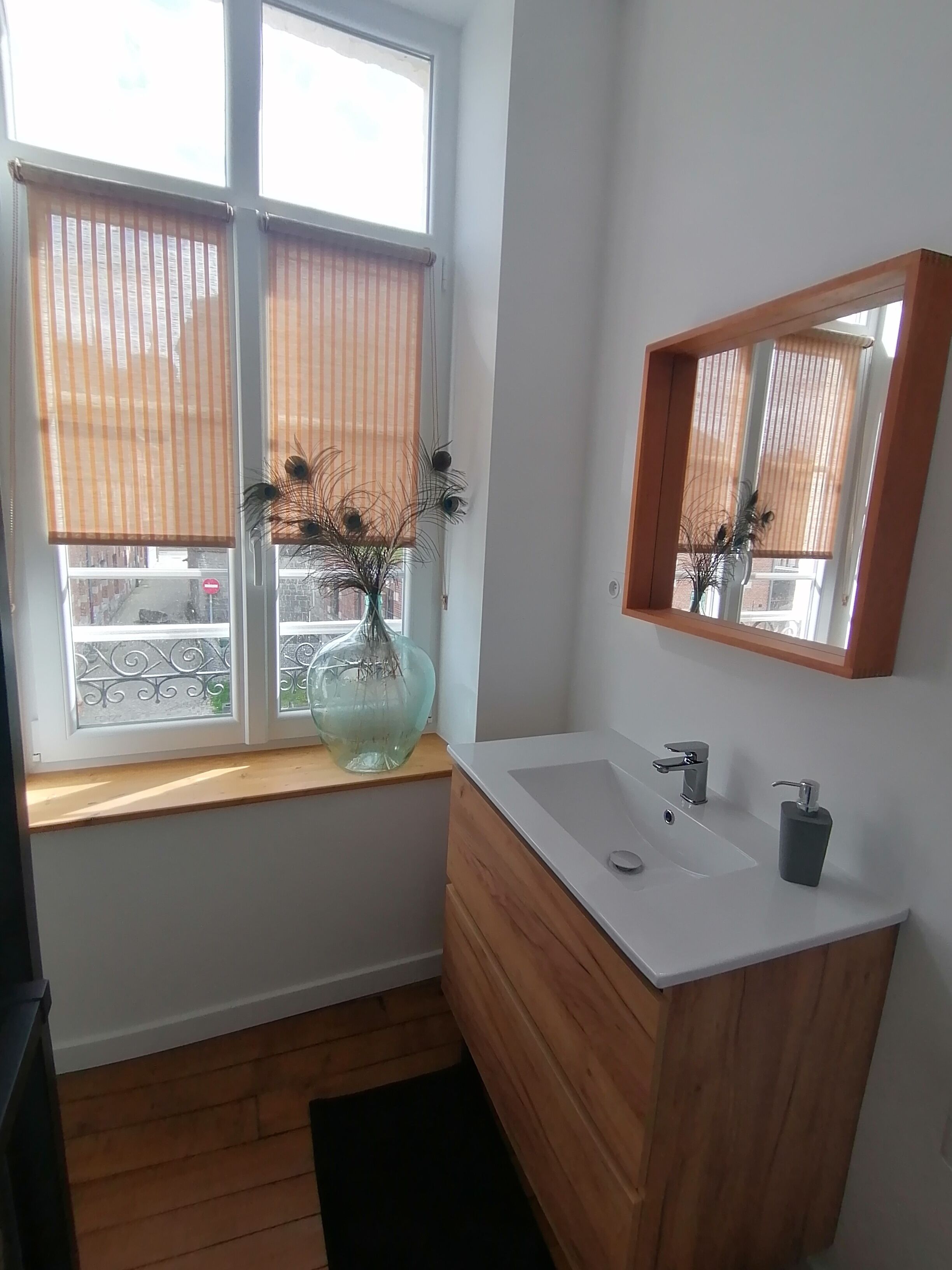Un baño con tocador de madera, un lavabo blanco y un espejo. Hay una ventana con persianas y un florero con plantas secas en el alféizar.