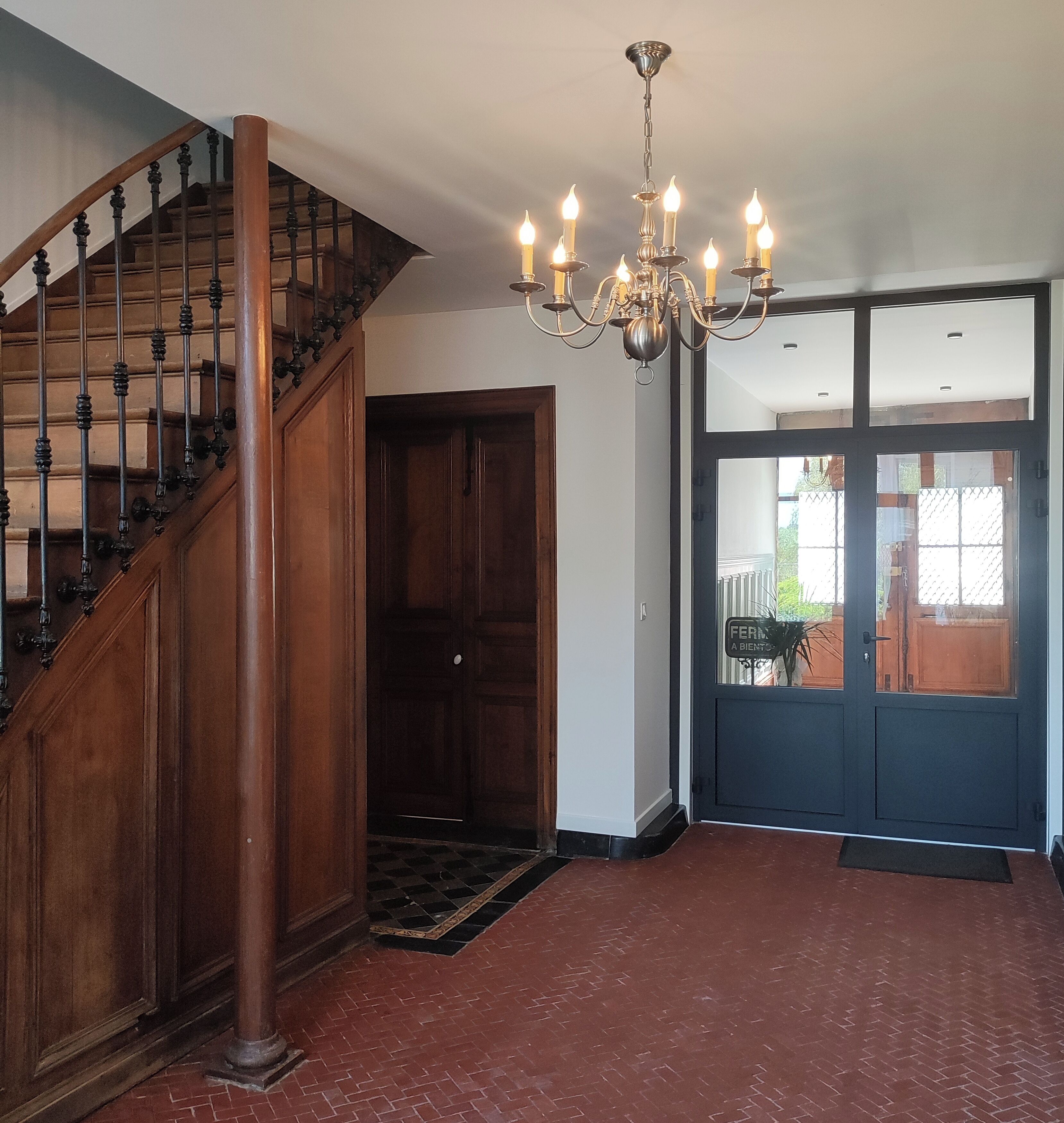 Un escalier en bois, avec une rampe noire, mène à une porte en bois sombre. Un lustre est suspendu au plafond, et un sol en briques rouges s’étend jusqu’à une porte en verre donnant sur l’extérieur.