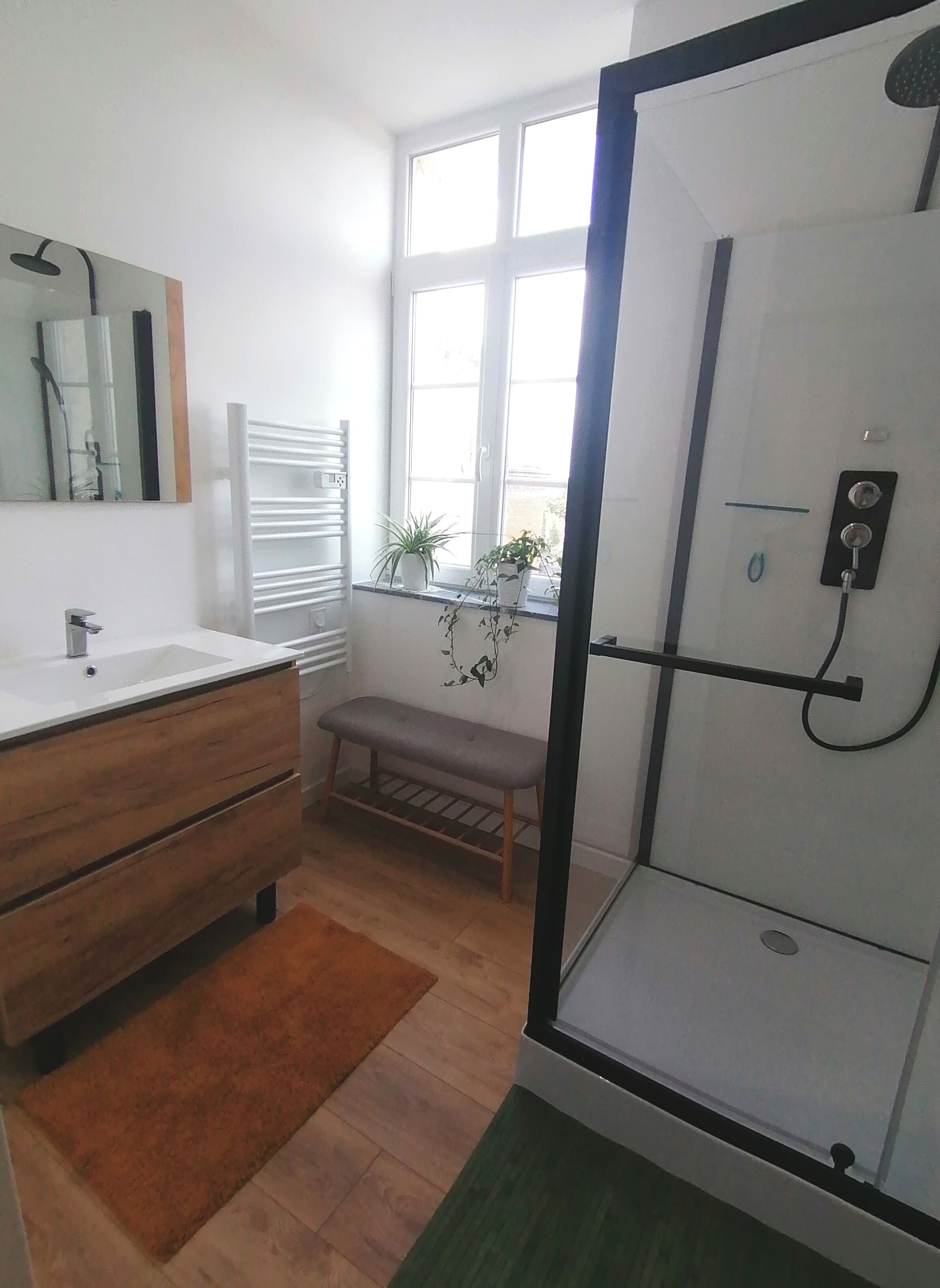 A bathroom with a shower, a sink, a mirror, a bench, and two potted plants.