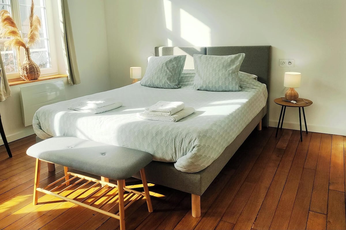 A neatly made bed with pillows and folded towels, a bedside table with a lamp, and a bench in a room with wooden flooring and a window with curtains.