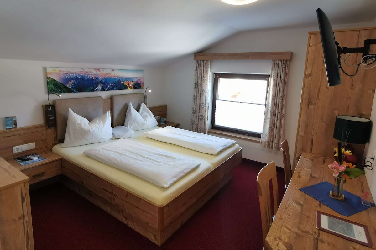 A bedroom with a wooden bed, a desk, a chair, a window with curtains, a painting on the wall, and a ceiling light.